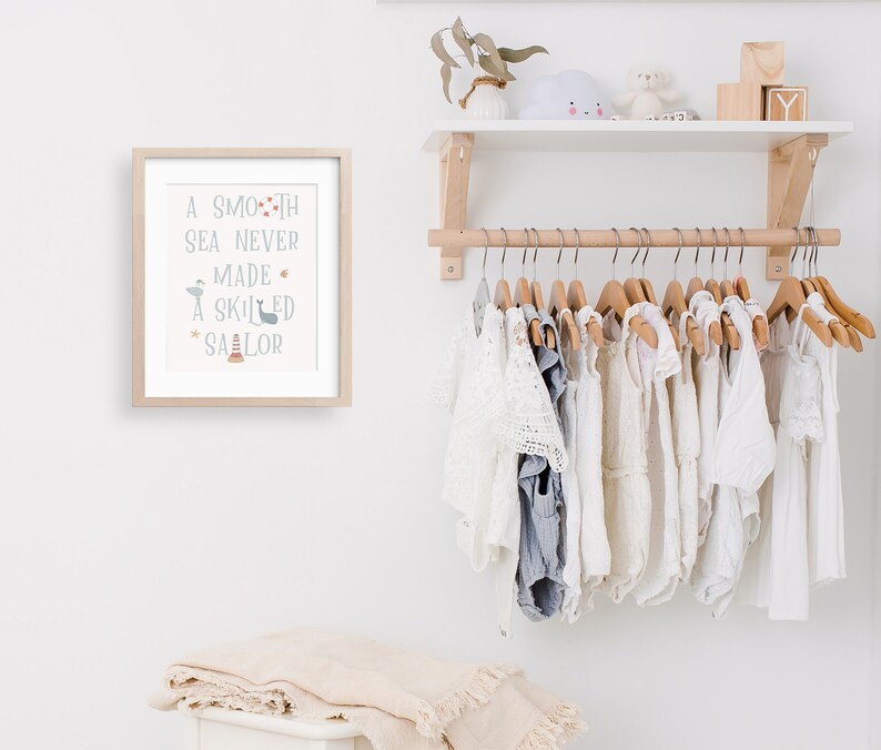 May include: A framed print with the text "A smooth sea never made a skilled sailor" with a nautical theme. The print is hanging on a white wall above a wooden shelf with a white surface. The shelf is holding a white cloud-shaped stuffed toy, a small wooden box, and a wooden block with the letter "Y" on it. Below the shelf is a wooden rod with a row of white clothing hanging on wooden hangers.
