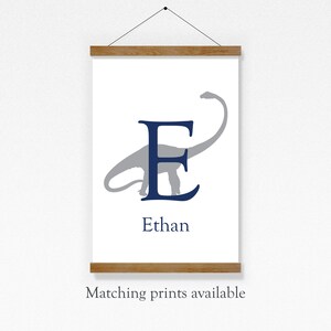 May include: A white canvas print with a blue letter E and a gray dinosaur silhouette. The name Ethan is printed below the letter. The print is hanging on a wall with a wooden frame.