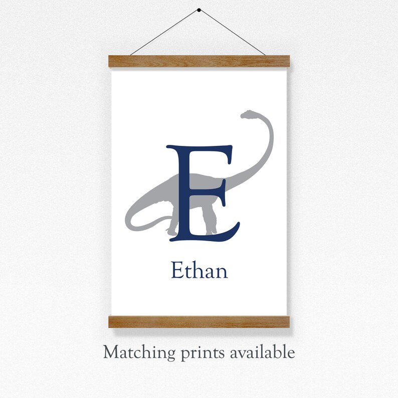 May include: A white canvas print with a blue letter E and a gray dinosaur silhouette. The name Ethan is printed below the letter. The print is hanging on a wall with a wooden frame.