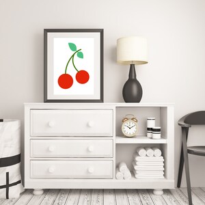 May include: White dresser with three drawers and a shelf above. The shelf has a gold alarm clock, two white jars, and white towels. A framed print of two red cherries with green stems is above the dresser. A black lamp with a white shade is on the dresser.