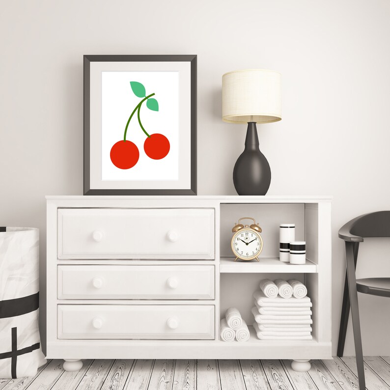 May include: White dresser with three drawers and a shelf above. The shelf has a gold alarm clock, two white jars, and white towels. A framed print of two red cherries with green stems is above the dresser. A black lamp with a white shade is on the dresser.