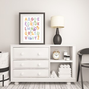 May include: A white dresser with three drawers and an open shelf. The dresser has a framed print of the alphabet with sea creatures. A gold alarm clock, a white lamp with a beige shade, and two white cylindrical containers are on the shelf. A white towel is folded on the shelf.