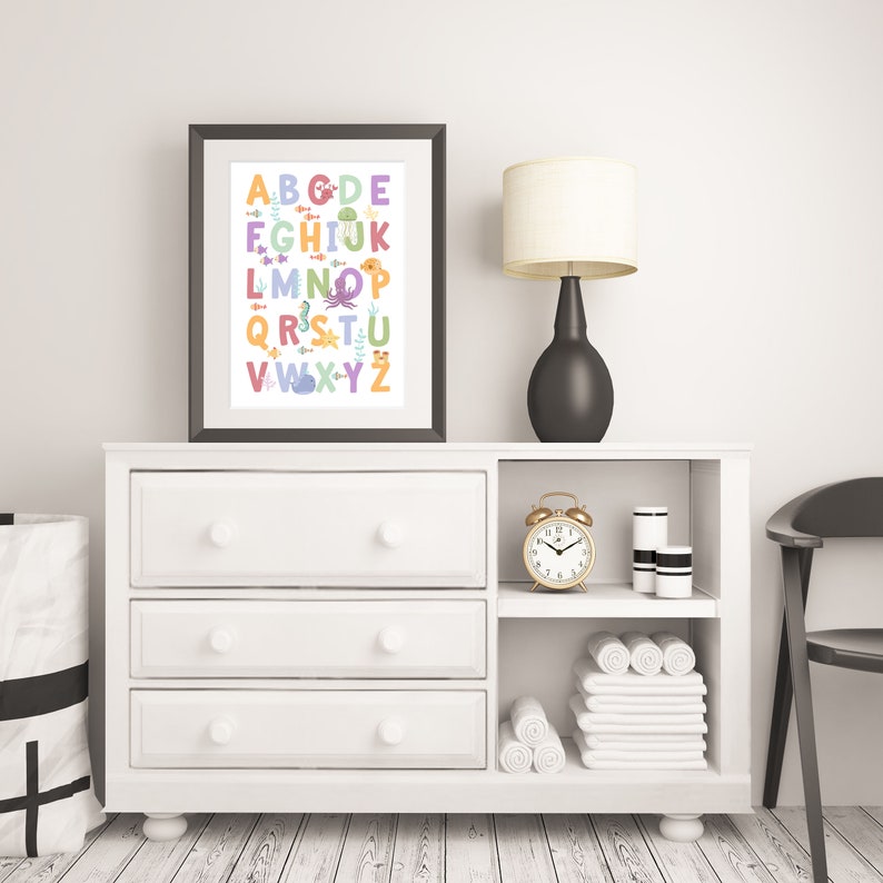 May include: A white dresser with three drawers and an open shelf. The dresser has a framed print of the alphabet with sea creatures. A gold alarm clock, a white lamp with a beige shade, and two white cylindrical containers are on the shelf. A white towel is folded on the shelf.