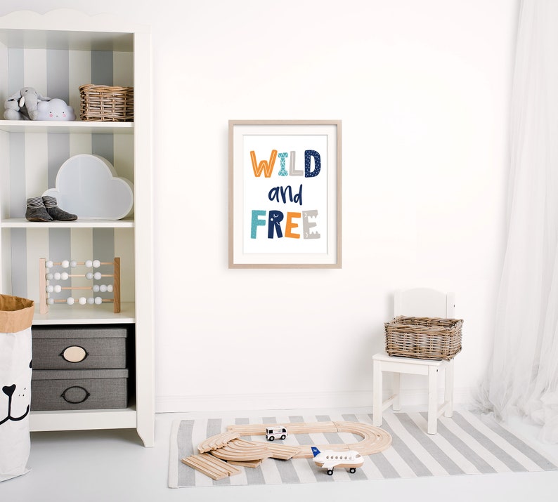 May include: A white framed print with the text "WILD and FREE" in colorful letters on a white wall in a nursery. The print is above a white wooden chair with a wicker basket.