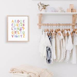 May include: A framed alphabet print with colorful letters and illustrations of sea creatures. The print is hanging on a white wall above a wooden clothes rack with white baby clothes hanging on it.