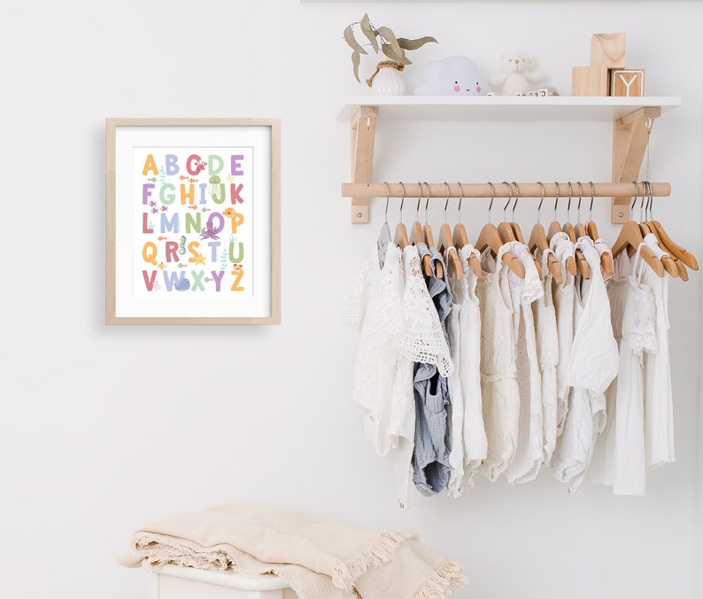 May include: A framed alphabet print with colorful letters and illustrations of sea creatures. The print is hanging on a white wall above a wooden clothes rack with white baby clothes hanging on it.