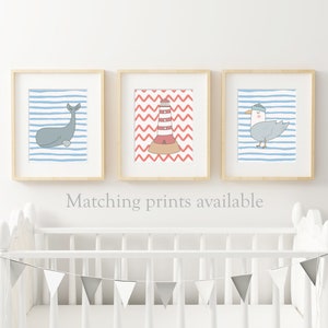 May include: Three framed prints with nautical themes. The first print features a whale on a blue and white striped background. The second print features a lighthouse on a red and white chevron background. The third print features a seagull on a blue and white striped background. The prints are displayed above a white crib with a gray and white bunting.
