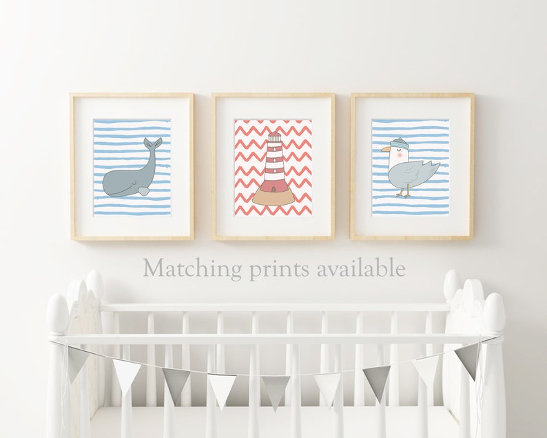 May include: Three framed prints with nautical themes. The first print features a whale on a blue and white striped background. The second print features a lighthouse on a red and white chevron background. The third print features a seagull on a blue and white striped background. The prints are displayed above a white crib with a gray and white bunting.