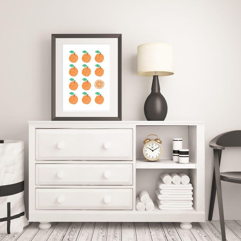 May include: A white dresser with three drawers and open shelves. A black framed print with a pattern of orange slices hangs above the dresser. A black lamp with a white shade sits on the dresser. A gold alarm clock sits on the shelf. White towels are folded on the shelves.