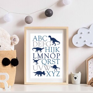 May include: A framed print with a white background and blue and gray text. The text is the alphabet with a dinosaur silhouette next to each letter. The frame is light wood.