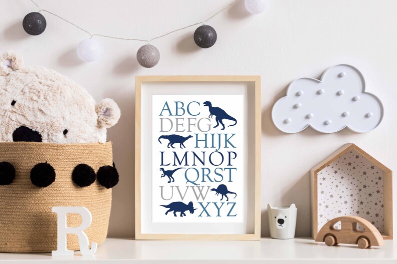 May include: A framed print with a white background and blue and gray text. The text is the alphabet with a dinosaur silhouette next to each letter. The frame is light wood.