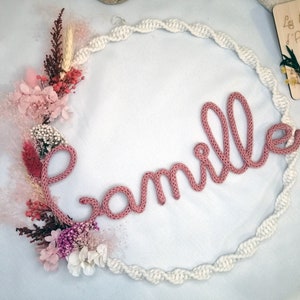 Peut inclure: Une couronne de fleurs séchées en rose, blanc et rouge est ornée d'un prénom en tricot rose "Camille". La couronne est faite d'une corde blanche en forme de cercle.