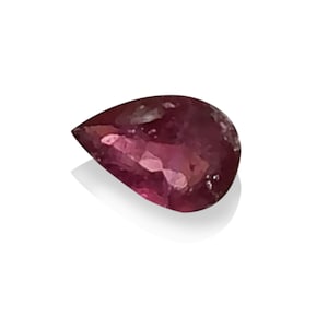 May include: A teardrop-shaped, faceted gemstone in a deep reddish-purple colour. The gem sits on a white background, with a soft shadow. The polished surface reflects light, highlighting its clarity and brilliance. Ideal for jewellery.