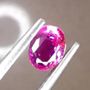 May include: A vibrant pink oval-shaped gemstone, likely a ruby, is held by tweezers. The gemstone is translucent and has a slight sparkle.