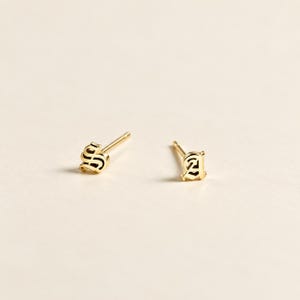May include: A pair of gold stud earrings featuring the letters S and A in an old English gothic font. Each earring has a thin post extending from the back of the letter, designed for piercing. The metal has a polished, reflective yellow gold finish.