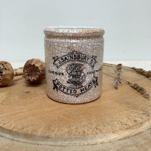 May include: A vintage J. Sainsbury's Potted Meats jar. The ceramic jar has a cracked, off-white glaze with brown lines. The label features black text and a logo. The jar is cylindrical with a slightly flared rim. The words "Superior" and "Home Made" are visible.