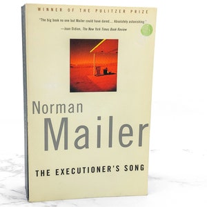 May include: A book cover with a yellow background and the title "THE EXECUTIONER'S SONG" in black text. The author's name, "Norman Mailer", is in gray text. The book cover also features a photograph of a gas station in the desert.