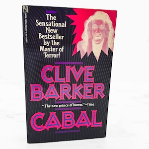 May include: A paperback book titled Cabal by Clive Barker with a black cover featuring thin white vertical lines. The cover displays a white illustration of a person with hands on their head against a pink burst, with text in pink and blue.
