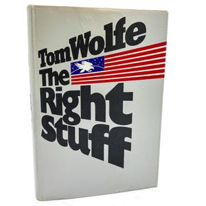 May include: A hardcover book titled The Right Stuff by Tom Wolfe with a light gray cover. The text is printed in large black letters. A graphic of a blue rectangle with a white eagle and red and white horizontal stripes appears on the front.
