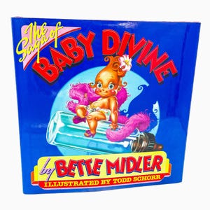 May include: A hardcover book titled "The Saga of Baby Divine" by Bette Midler, illustrated by Todd Schorr. The cover features a cartoon baby with orange hair, pink boa, and a flower, sitting on a baby bottle against a blue background.
