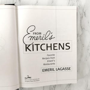 以下が含まれることがあります： 「From Emeril's Kitchens」というタイトルがエレガントな筆記体と太字のブロック文字で書かれた、開かれた料理本のページ。その下に、エメリル・ラガッセ著「Favorite Recipes from Emeril's Restaurants」と書かれています。ウィリアム・モローのインプリントが見えます。