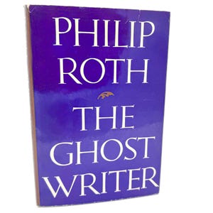 May include: A hardcover book titled The Ghost Writer by Philip Roth with a deep purple cover. The title and author name are printed in large white serif capital letters. A small gold leaf-shaped graphic is centered between the author and title.