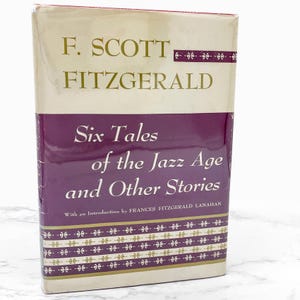 May include: A hardcover book titled "Six Tales of the Jazz Age and Other Stories" by F. Scott Fitzgerald. The book has a cream-colored top and bottom with a deep purple center section. Gold lettering and decorative elements are present.
