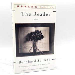 May include: A hardcover book titled "The Reader" by Bernhard Schlink. The cover is white with black text and features a black and white image of an open book with a bouquet of flowers. The book is part of Oprah's Book Club.