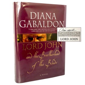 May include: A hardcover book titled Lord John and the Brotherhood of the Blade by Diana Gabaldon. The maroon cover features a close-up of a person's eyes and gold lettering. A separate white rectangular label displays a signature and the text LORD JOHN.