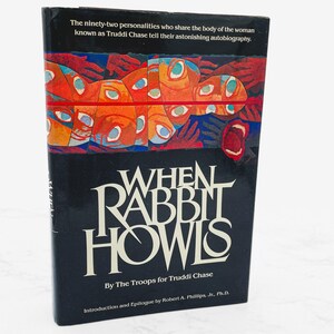 May include: A hardcover book titled "When Rabbit Howls" with a black cover. The cover features the title in large white letters and an illustration of faces in orange, red, and blue. The text at the top reads, "The ninety-two personalities who share the body of the woman known as Truddi Chase tell their astonishing autobiography."