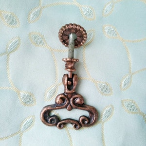 May include: A decorative, ornate, copper-toned cabinet knob with a floral design. The knob has a round, metal base and a screw-on attachment.