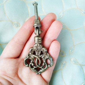 May include: A vintage ornate metal drawer pull with a decorative scroll design. The pull is finished in a brass tone and has a screw for attachment.
