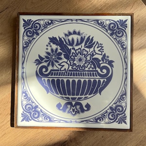 Może przedstawiać: Kwadratowa płytka ceramiczna z niebieskim kwiatowym wzorem w białym okręgu, osadzona w drewnianej ramie. Kwiatowy wzór przedstawia wazon wypełniony kwiatami i dekoracyjnymi zawijasami na obramowaniu. Płytka leży na drewnianej powierzchni.