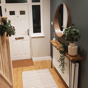 May include: An inviting hallway with a wooden console table, a round wooden mirror, and a white radiator. The table has black hairpin legs. A striped rug and a white door with glass panels are also visible.