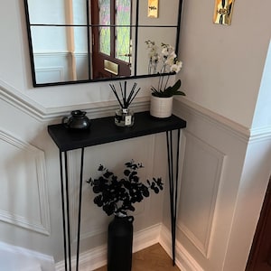 K&ouml;nnte beinhalten: Ein schwarzer Konsolentisch mit Haarnadelbeinen, auf dem eine schwarze Keramikvase mit einem Strau&szlig; schwarzer Eukalyptus&auml;ste, ein schwarzer Keramik Krug und ein Glasdiffusor mit schwarzen Schilfrohren stehen. Der Tisch steht in einem Flur mit wei&szlig;en W&auml;nden und einem schwarz gerahmten Spiegel mit einem Gittermuster.