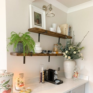 Könnte beinhalten: Ein rustikales Holzregal mit zwei Ebenen, auf dem eine weiße Keramikvase mit einem Strauß weißer und grüner Blumen, ein gerahmtes Bild und eine Kaffeemaschine stehen. Die Regale sind an einer weißen Wand mit einer Leuchte darüber befestigt.