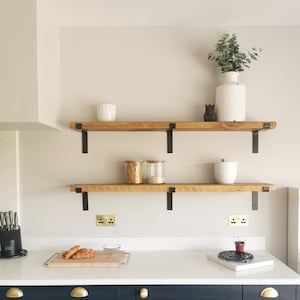 May include: Two wooden shelves with black metal brackets mounted on a white wall above a dark blue kitchen cabinet with brass hardware. The shelves are decorated with white ceramic vases, glass jars, and a small potted plant.