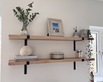 May include: Two wooden floating shelves with black metal brackets are mounted on a white wall. The top shelf holds a framed picture, a glass vase with greenery, and a small concrete pot. The bottom shelf displays books, a decorative sphere, and a candle.