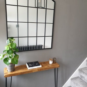 Op de afbeelding: Een moderne entree met een zwarte spiegel met raster, een houten console met zwarte haarspeldpoten en een potplant. Een boek en kaarsen staan op de tafel. De muren zijn neutraal grijs geschilderd.