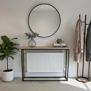 May include: A contemporary hallway arrangement with a circular black mirror, a console table with a light wood surface, and a coat stand. A potted plant and decorative items are also present. Two coats hang on the stand.