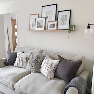 May include: A light gray sofa with a variety of throw pillows, including a fluffy gray pillow, a patterned pillow with gray and white stripes, and a solid gray pillow. The sofa is in front of a white wall with a shelf holding black and white framed artwork.
