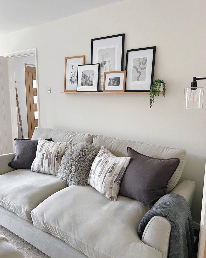 May include: A light gray sofa with a variety of throw pillows, including a fluffy gray pillow, a patterned pillow with gray and white stripes, and a solid gray pillow. The sofa is in front of a white wall with a shelf holding black and white framed artwork.