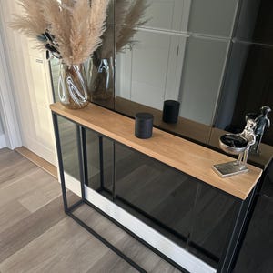 May include: A contemporary console table with a black metal frame and a light wood top. It has a glass front and is adorned with a vase of pampas grass, a black cylindrical object, and a small silver sculpture.