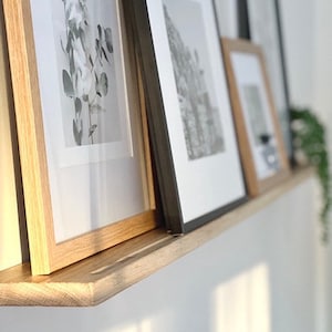 May include: A wooden shelf with three framed pictures. The shelf is mounted on a white wall. The pictures are in black and white and feature different subjects. The shelf is made of natural wood and has a simple design.