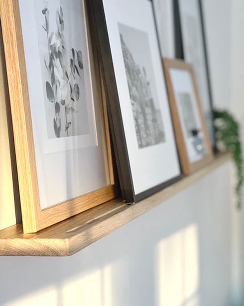 May include: A wooden shelf with three framed pictures. The shelf is mounted on a white wall. The pictures are in black and white and feature different subjects. The shelf is made of natural wood and has a simple design.