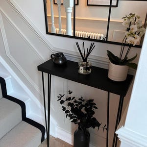 K&ouml;nnte beinhalten: Schwarzer Konsolentisch aus Metall mit einer schwarzen Keramikvase und schwarzen Blumenstielen. Der Tisch hat d&uuml;nne Haarnadelbeine und einen schwarzen Keramik Krug auf der oberen Ablage. Eine klare Glasflasche mit schwarzen Schilfrohren befindet sich ebenfalls auf der oberen Ablage.