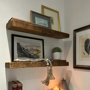 Könnte beinhalten: Zwei Holzregale mit rustikalem Finish, montiert an einer weißen Wand. Das obere Regal hat einen gerahmten Druck eines Schwans und einen gerahmten Druck einer Landschaft. Das untere Regal hat einen gerahmten Druck einer Landschaft, eine kleine Topfpflanze und eine Schnur aus Holzperlen mit Quasten.