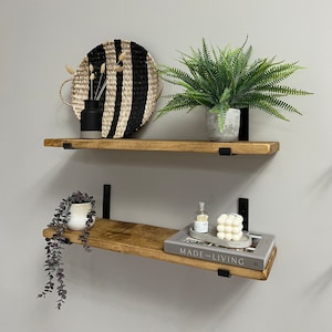 Pode incluir: Duas prateleiras de madeira com suportes de metal preto, decoradas com plantas, uma bandeja tecida em preto e branco, um vaso pequeno, um livro intitulado "Made for Living", uma garrafa de vidro e bolas decorativas brancas.