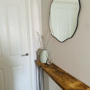 May include: A rustic wooden console table with black hairpin legs, a white vase with willow branches, and a diffuser bottle on top. The table is placed in a hallway with a white door and a black, irregular-shaped mirror on the wall.