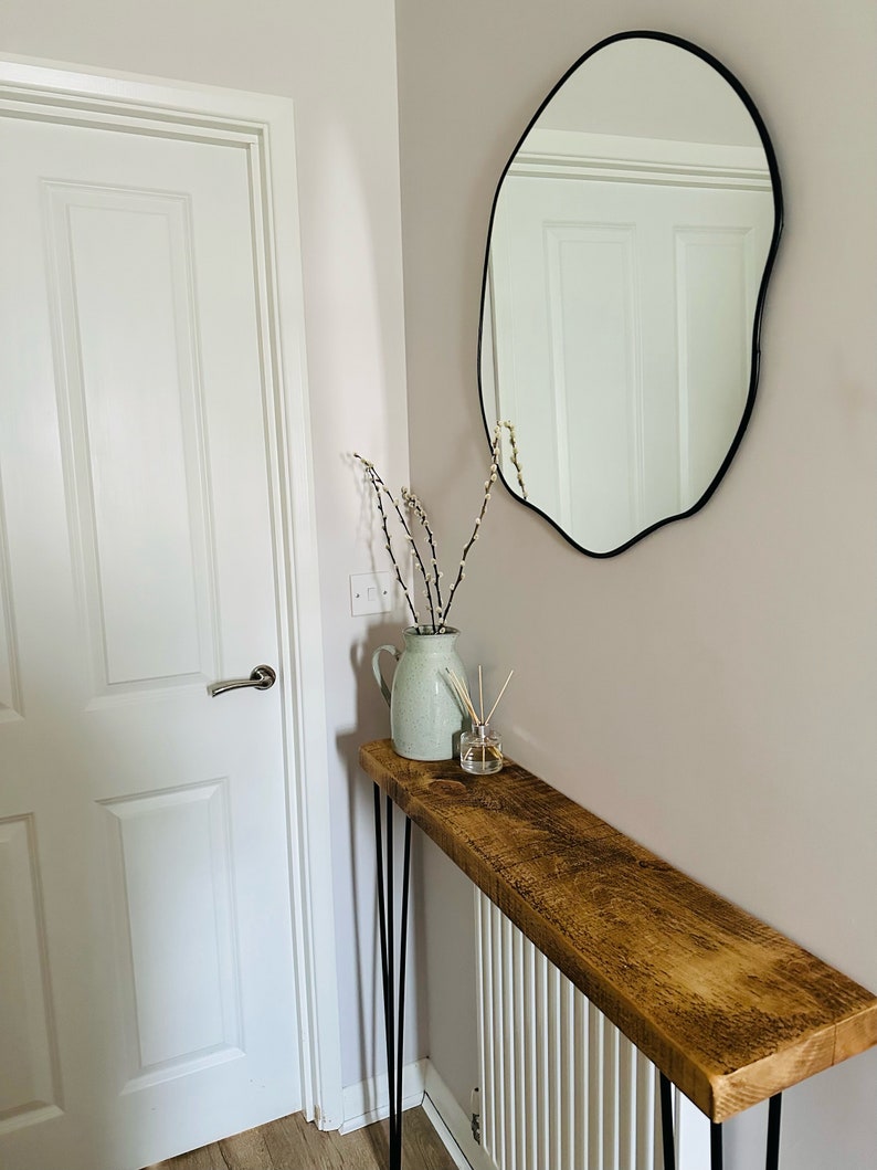 May include: A rustic wooden console table with black hairpin legs, a white vase with willow branches, and a diffuser bottle on top. The table is placed in a hallway with a white door and a black, irregular-shaped mirror on the wall.